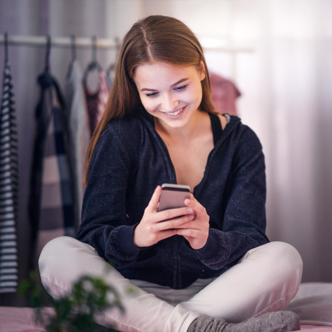 Happy teenage girl looking at smartphone, relaxing at home, enjoy using mobile online apps, playing games on mobile phone, checking messages or social media messages. Soft selective focus. Free pregnancy test near me
