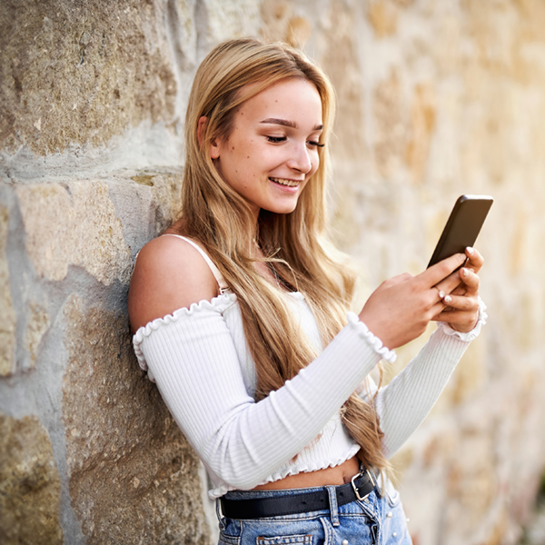 Cheerful teenager student girl standing in front of a vintage stone wall and orering something online with a smart phone - Young woman checking her social media feed and sharing a story