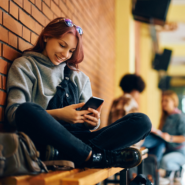 Happy teenage girl using smart phone while relaxing in high school hallway.