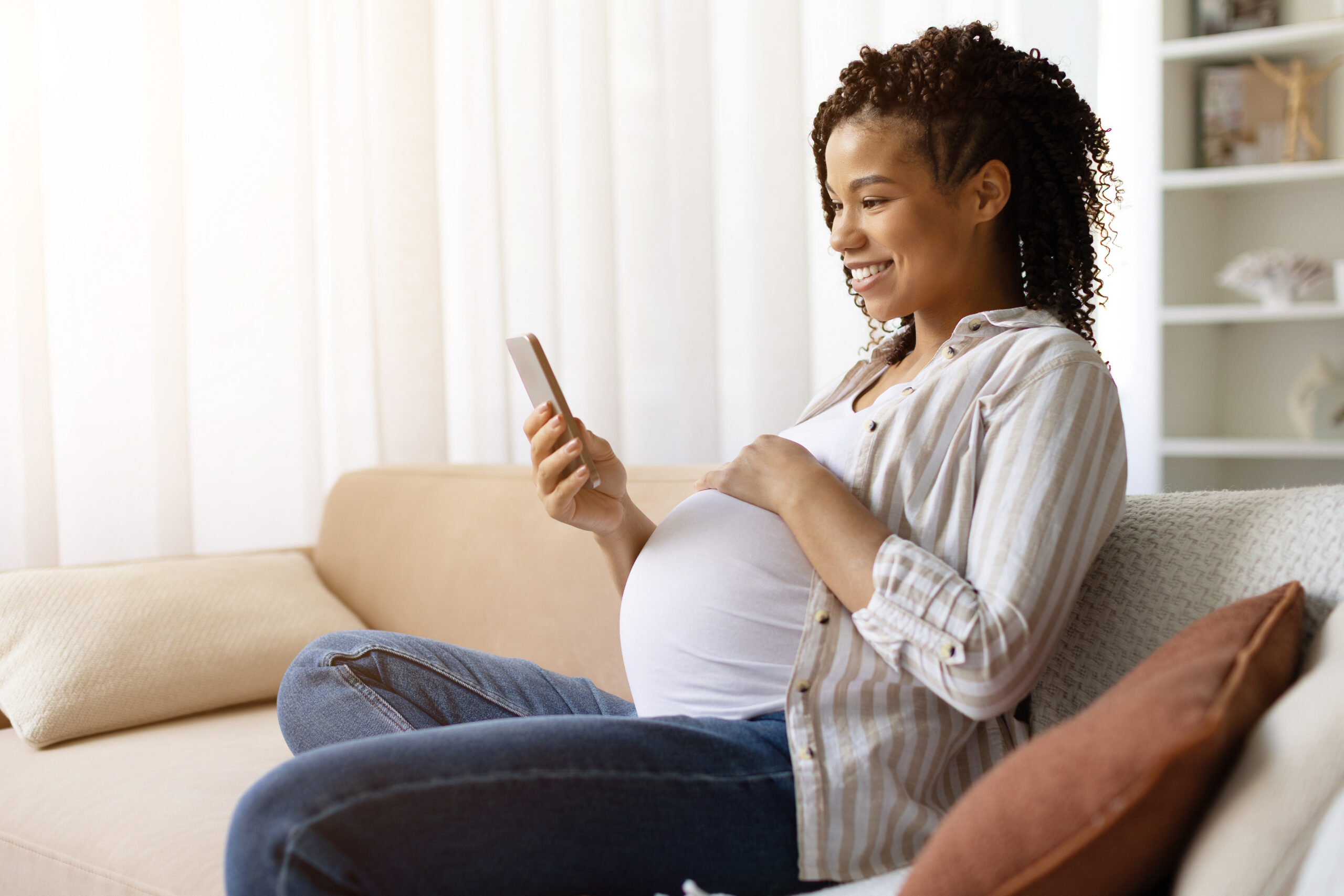Smiling African American pregnant woman relaxing on sofa and looking at smartphone